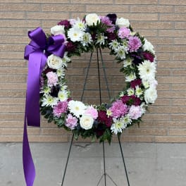 Circular floral wreath with white, pink, and purple blooms on a stand with a purple ribbon