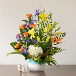 Tall mixed bouquet with yellow lilies, blue irises, orange roses, and white hydrangea in a round vase