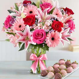 Pink and red mixed bouquet in a glass vase with a pink ribbon, beside chocolate-covered strawberries