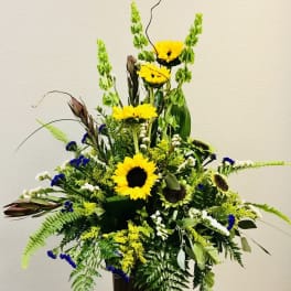 Tall arrangement of yellow sunflowers, green bells of Ireland, and blue accents in a dark rectangular container