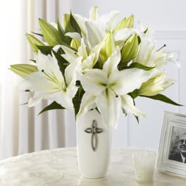 White lily arrangement in a tall white vase with a silver cross emblem on a light table.