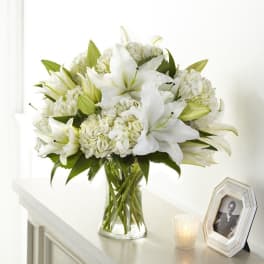White lilies and hydrangeas arranged in a clear glass vase beside a framed photo and small candle.