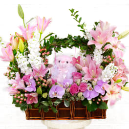 Pink lilies and orchids arranged in a wicker basket with a teddy bear