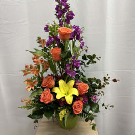 Tall mixed flower arrangement with orange roses, purple blooms, and a yellow lily in a vase