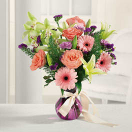 Mixed bouquet of pink gerbera daisies, roses, lilies and green orchids in a purple vase with cream ribbon