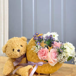 Teddy bear beside a crescent flower arrangement in a gold container