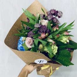Handheld bouquet of purple, pink, blue, and green flowers wrapped in brown paper