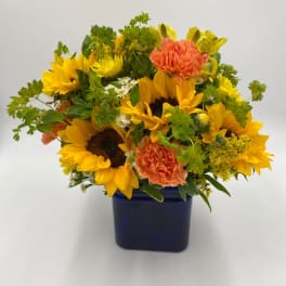 Low arrangement of yellow sunflowers and orange carnations in a blue cube vase