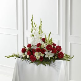 Red and white sympathy spray with roses, gerbera daisies, lilies, and tall white blooms on a draped table.