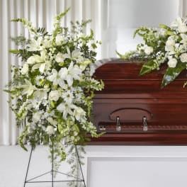 Standing spray of white lilies, roses, and daisies beside a wood casket in a funeral setting