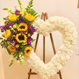 Heart-shaped white floral wreath on an easel beside a colorful sunflower bouquet.