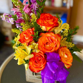 Bouquet of orange roses and purple flowers in a wooden container with a purple ribbon