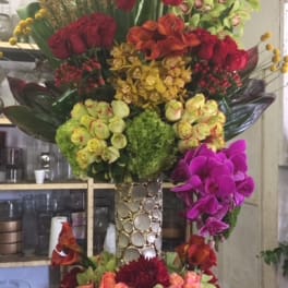 Tall floral arrangement with red, orange, yellow, green, and purple blooms in a decorative vase.