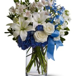 White lilies and roses with blue and white blooms in a clear glass vase with a blue ribbon bow