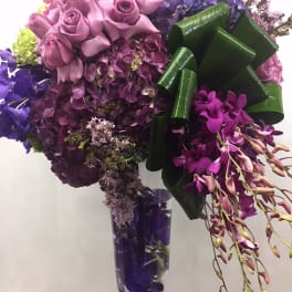 Purple roses and orchids arranged in a clear vase with a large green ribbon