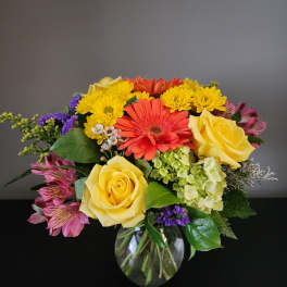 Mixed bouquet of yellow roses, gerbera daisies, and purple flowers in a glass vase