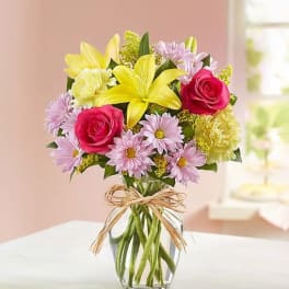 Bouquet of yellow lilies, pink roses, and lavender daisies in a glass vase