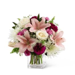 Pink lilies, white roses, and burgundy carnations arranged in a clear glass cube vase