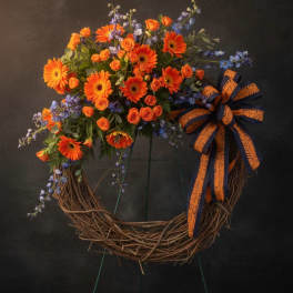 Grapevine wreath on stand with orange gerbera daisies, orange roses, blue delphinium and a large orange and navy bow