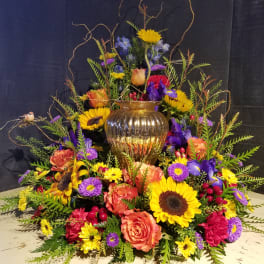 Large mixed floral arrangement with sunflowers, roses, and purple blooms in a gold vase