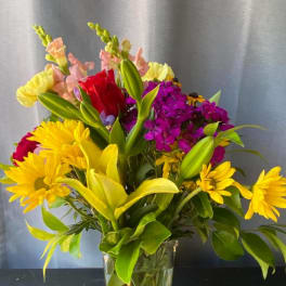 Mixed bouquet of yellow daisies, lilies, and purple flowers in a clear vase