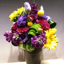 Tall mixed arrangement of yellow sunflowers, purple lisianthus, and magenta mums in a dark vase
