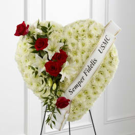 Heart-shaped white mum funeral spray with red roses, white lilies, and a tribute ribbon on an easel