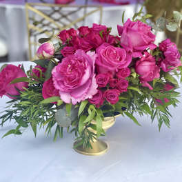 Low centerpiece of bright pink roses and peonies with greenery in a gold compote vase on a white table
