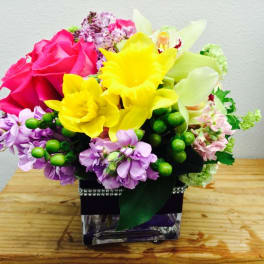 Bright mixed bouquet with pink roses, yellow daffodils, and purple flowers in a square vase