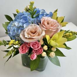 Bouquet of blue hydrangeas, peach roses, pink spray roses, and yellow lilies in a glass vase