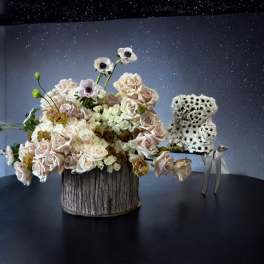 Blush roses and white anemones arranged in a rustic wood container beside a small white flower bear.
