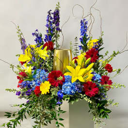 Tall floral arrangement with yellow lilies, red daisies, and blue flowers in a gold vase