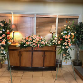 Three floral funeral sprays with peach and white flowers on stands and a casket spray on a cabinet