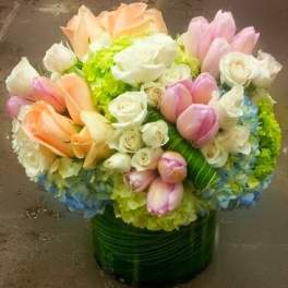 Round arrangement of peach and pink tulips, white roses, and hydrangeas in a leaf-wrapped glass vase