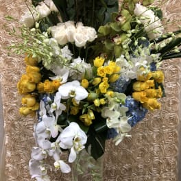Tall bouquet of yellow roses, white orchids, and blue hydrangeas in a clear vase