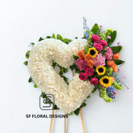 Heart-shaped white standing wreath with colorful roses, sunflowers, and mixed blooms on one side.