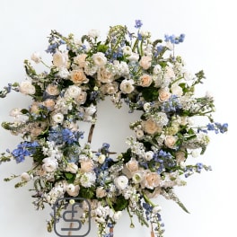 Large round wreath of cream roses and white flowers with soft blue accents on a wooden easel.