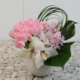 Compact arrangement of pink tulips, white orchids, and pastel hyacinths in a white textured vase with looping green blades.