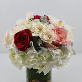 Compact arrangement of red and cream roses, pink rose, hydrangeas, and orchid in a glass cube vase