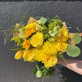 Hand-tied bouquet of bright yellow roses, mums, and tulips held against a gray wall.