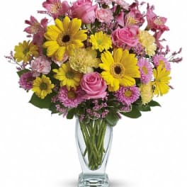 Mixed bouquet of yellow daisies, pink roses, and carnations arranged in a clear glass vase