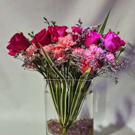 Tall arrangement of hot pink roses and pink carnations in a clear rectangular vase with striped foliage.