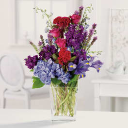 Tall mixed bouquet of purple, pink, and blue flowers in a clear glass vase