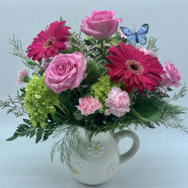 Pink roses, gerbera daisies, green hydrangea, and carnations in a white daisy pitcher with a blue butterfly pick.