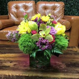 Low glass vase of yellow mums, purple blooms, green dianthus, and an orchid on a wooden table