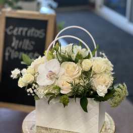 White floral arrangement in a handled gift box with roses and orchids