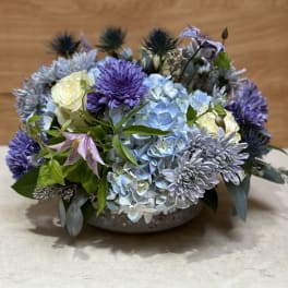 Low arrangement of blue hydrangeas, purple mums, and white roses in a shallow bowl