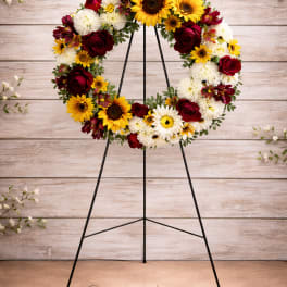 Circular wreath of sunflowers, red roses, and white daisies on a black stand
