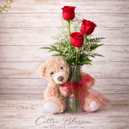 Three red roses in a glass vase with a teddy bear and red ribbon