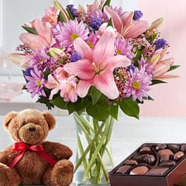 Pink lilies and daisies in a glass vase with a teddy bear and chocolates